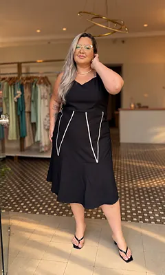 Mulher de óculos e cabelo longo com vestido midi preto de vivos brancos, na porta da Realce